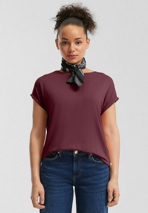 Junge Frau mit zurückgebundenen lockigen Haaren, die ein dunkelrotes Kurzarmshirt, dunkelblaue Jeans und ein schwarzes, gemustertes Halstuch trägt.