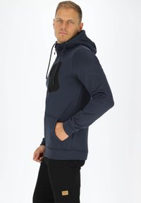 Felpa cappuccio blu navy con accenti neri, dotata di tasca a marsupio, zip e tessuto testurizzato. Il modello sta in piedi con le mani nelle tasche.