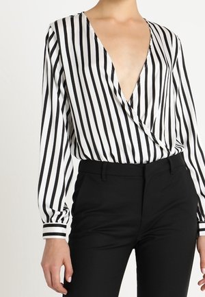 Kobieta w czarno-białej koszuli z pionowymi paskami i głębokim dekoltem w serek oraz czarnych spodniach z wysokim stanem, widoczne tułów i ramiona.