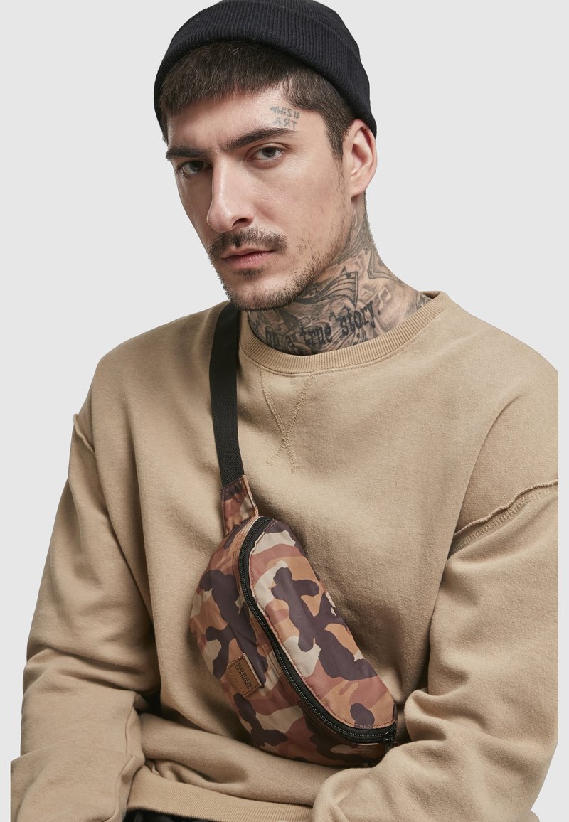 Homme tatoué portant un bonnet noir, un sweat-shirt beige et un sac bandoulière camouflage sur fond uni.