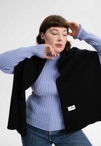 Zwarte sjaal over een lichtblauwe geribbelde coltrui. De outfit is gecombineerd met spijkerbroek. Een klein label op de sjaal zegt "MELA."