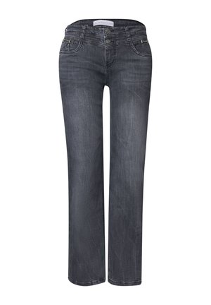 Femme portant un jean gris et une chemise rose clair à manches longues boutonnée avec les manches retroussées, debout devant un fond uni.