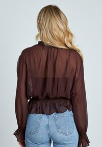 Blusa marrón transparente con cuello con volantes y cintura elástica, con mangas largas y puños con volantes, combinada con jeans de tiro alto de color azul claro.