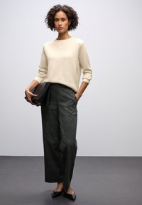 Donna con capelli ricci, che indossa un maglione color crema, pantaloni a gamba larga a quadri, ballerine nere e tiene una pochette nera su uno sfondo neutro.