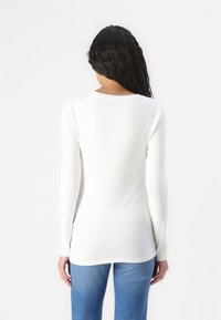Top bianco a maniche lunghe realizzato in tessuto leggero con una vestibilità rilassata, caratterizzato da un collo rotondo e una texture liscia. Abbinato a jeans blu.