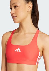 Punainen urheilu crop-top, joka on valmistettu joustavasta materiaalista, jossa on neliömäinen kaula-aukko ja valkoiset raidat sekä brändilogolla etuosassa.