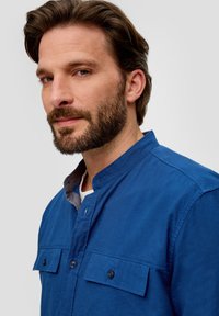 Blauwe overhemd met een Mandarin-kraag, gestructureerde stof, twee borstzakken en een contrasterende grijze onderkraag.