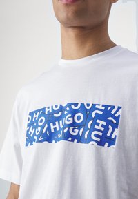 T-shirt en coton blanc avec un graphisme bleu affichant en répétition le texte "HUGO" en blanc. Col rond et manches courtes, tissu léger.