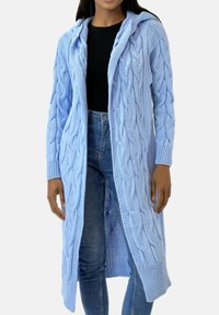 Jasnoniebieski dziergany sweter z warkoczowym wzorem, długimi rękawami i kapturem. Ma otwarty przód i prążkowane mankiety. Nałożony na czarny top.