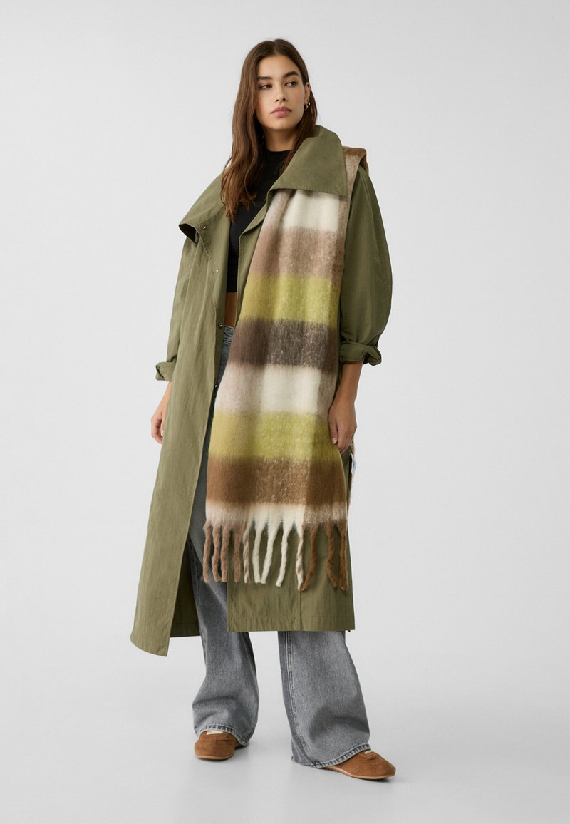 Cappotto verde oliva con una sciarpa grande e decorata che presenta fasce di colore marrone, crema e verde. La sciarpa ha bordi frangiati. Pantaloni grigi larghi.