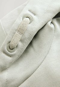 Pullover grigio chiaro con tessuto texturizzato, dotato di un grommet con logo e di un dettaglio decorativo con una cintura intrecciata. Finitura liscia e morbida.