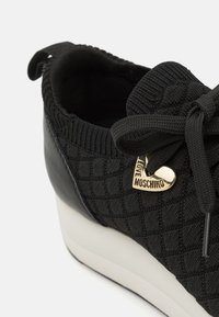 Baskets noires avec un upper en tricot texturé, des accents en cuir lisse, un lacet en ruban et un charm en forme de cœur doré avec "LOVE MOSCHINO."