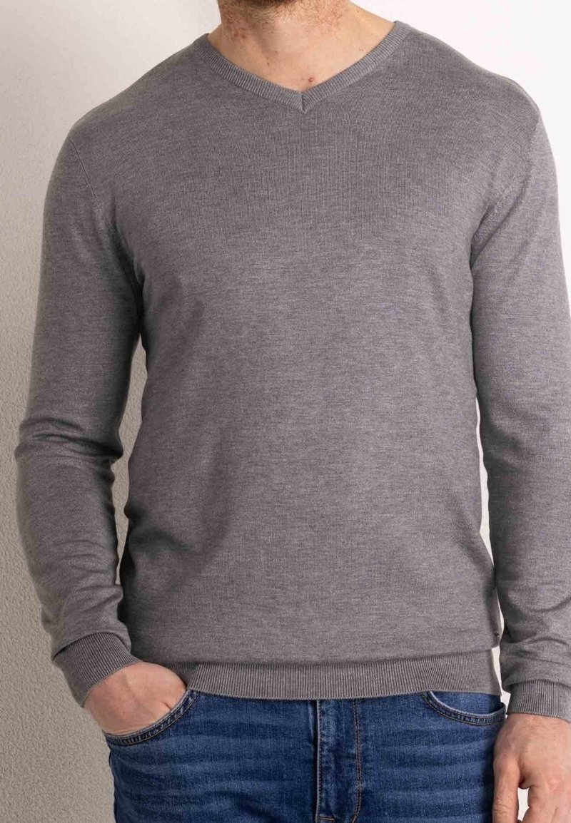 Pull gris à manches longues avec col en V, fabriqué en maille douce. Comprend des poignets et une ceinture côtelés. Porté avec un jean bleu.