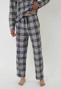 Man tilt een grijs en zwart geruit flanellen shirt omhoog om bijpassende geruite pyjamabroek met zwarte trekkoord te onthullen, blootsvoets op een witte achtergrond.
