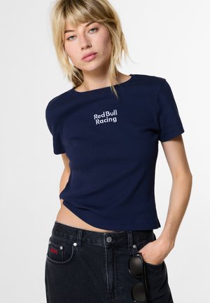 Jonge vrouw met blond haar die een donkerblauw Red Bull Racing t-shirt en zwarte spijkerbroek draagt, met een zonnebril die uit de zak hangt.