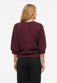 Femme aux cheveux blonds portant un haut bordeaux à manches bouffantes et un pantalon noir, debout de dos sur un fond blanc uni.