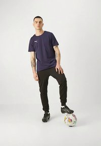 Mladý muž v tmavém sportovním oblečení stojící s jednou nohou na barevném fotbalovém míči na jednobarevném světlém pozadí.