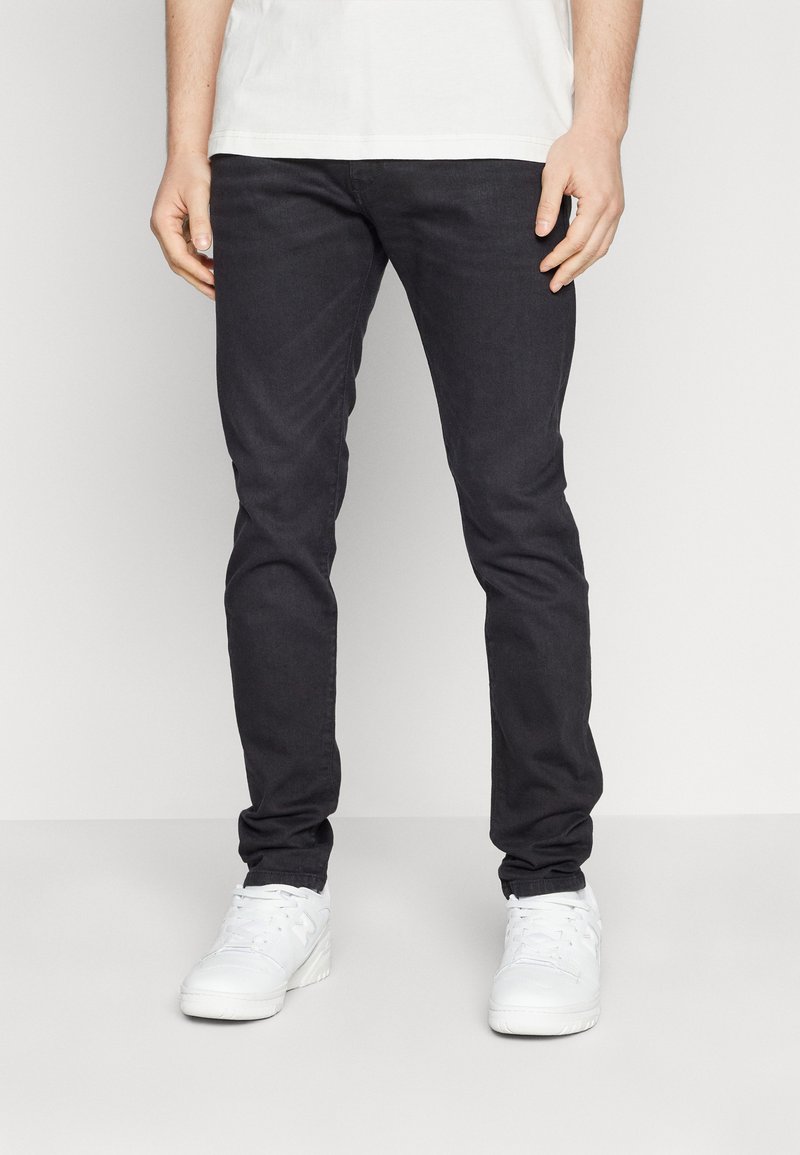 Svarta slim-fit jeans med en slät textur, avsmalnande ben och minimala sömdetaljer, i kombination med vita sneakers.