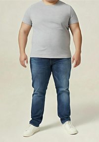 Adulte portant un t-shirt gris uni à manches courtes, un jean bleu et des baskets blanches, debout devant un fond beige neutre.