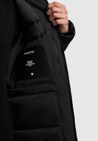 Schwarze isolierte Jacke mit einer glatten, glänzenden Oberfläche. Innen befindet sich ein Etikett mit dem Markennamen, dem Gründungsjahr und dem Logo.