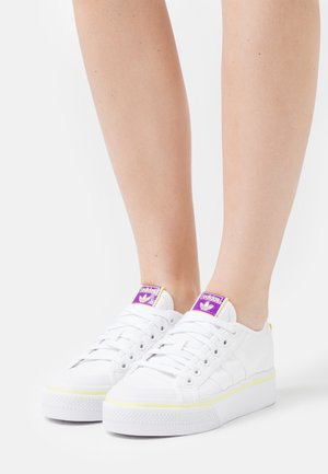 Zapatillas deportivas de lona blanca con una suela gruesa blanca y un acento amarillo, con marca púrpura en la lengüeta y un acabado texturizado.