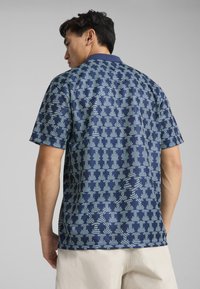 La chemise à manches courtes présente un fond bleu marine avec un motif géométrique bleu clair. Fabriquée en tissu respirant, elle comprend un col classique et une coupe décontractée.