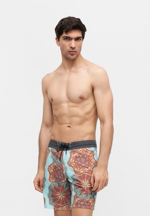 Νέος άνδρας με σκουρόχρωμα μαλλιά που φοράει μαγιό με κόκκινα και μπλε γεωμετρικά σχέδια, στέκεται σε απλό ανοιχτόχρωμο φόντο.