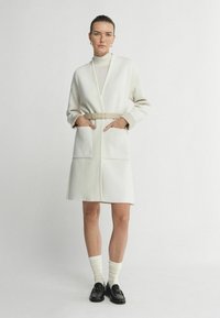 Lang hvid kjole lavet af blødt materiale, med et bælte, store lommer og en afslappet pasform. Modellen bærer cremefarvede sokker og loafers.