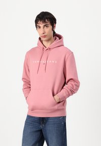 Felpa rosa in cotone con tasca frontale, logo bianco “TOMMYJEANS”, cappuccio regolabile con cordini e polsini e orlo a costine. Indossata con jeans blu.