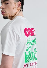 T-shirt de algodão branca com grande texto em rosa "OBEY" e gráfico floral verde nas costas, mangas curtas e decote redondo.