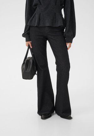 Kvinde iført sorte vide bukser og mørkegrå peplum top, der holder en sort lædertaske, står på en hvid baggrund.