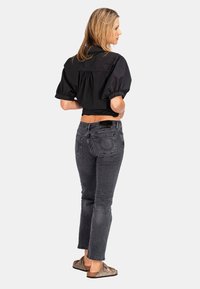 Schwarzes cropped Shirt mit puffigen Ärmeln, kombiniert mit dunkelgrauen, eng anliegenden Jeans. Das Modell trägt braune Slipper. Schlicher Hintergrund.