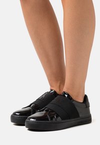 Black patent leather sneakers featuring wide elastic straps and a textured sole, with branding on the side and sleek minimalist design.