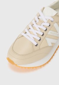 Baskets beiges avec des accents blancs, tige en cuir lisse, bout rond, semelle blanche épaisse avec contour orange, et lacets blancs passés dans des œillets en argent.