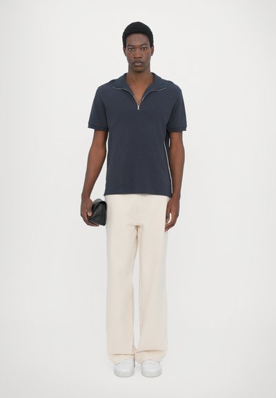 Navy zippered polo shirt with short sleeves, paired with cream wide-leg pants and white sneakers. Model holds a black clutch.