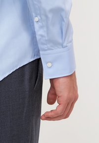 Cămașă albastră deschis cu mâneci lungi, având o textură netedă, cu manșete cu buton și o croială ajustată. Purtați-o cu pantaloni texturați negri.