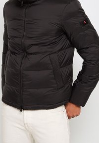 Svart vadderad jacka med framslits, sidofickor och liten logotyp på vänster ärm, bärs med vita byxor.