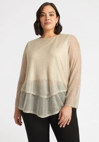 Top beige trasparente con maniche lunghe, design stratificato, motivo floreale e dettagli luccicanti, indossato sopra pantaloni scuri.