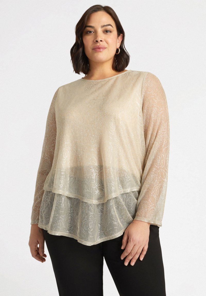 Top beige trasparente con maniche lunghe, design stratificato, motivo floreale e dettagli luccicanti, indossato sopra pantaloni scuri.