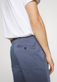 Modré bavlněné chinos kalhoty mají zadní kapsu na knoflík a malý logo akcent. Látka vypadá hladce a má mírný lesk.