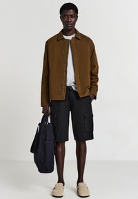 Mann trägt eine braune Jacke, ein hellgraues Hemd, schwarze Cargo-Shorts, beige Slipper, hält eine dunkelblaue Tragetasche und steht vor einem einfarbigen Hintergrund.