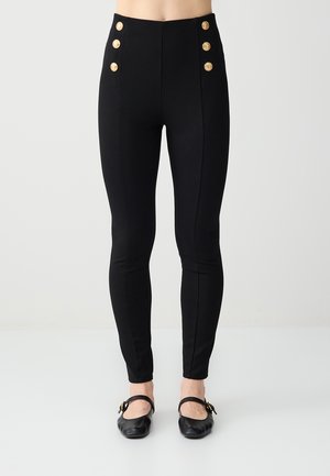 Pantalon noir slim taille haute avec six boutons décoratifs dorés à la taille, porté avec des chaussures plates Mary Jane noires.