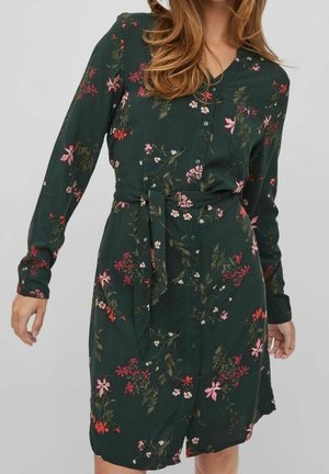 Vihreä kukkamekko pitkillä hihoilla, napillinen edusta, vyötäröllä vyö ja vaalealle kankaalle painettuja pinkkejä ja valkoisia kukkakuvioita.