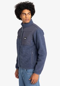 Fleece jacket in navy blue with a soft texture, zip pockets, and a contrasting material panel at the chest. Features a high collar and zip front.
