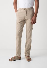 Beige linen pants with an elastic waistband and drawstring, featuring side pockets, styled with beige open-toed sandals.