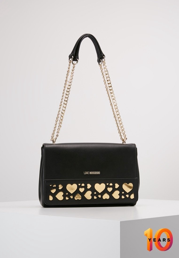 Sac à main en cuir noir avec des découpes en forme de cœur dorées à l'avant, doté d'une chaîne dorée et de détails de logo. Forme rectangulaire compacte.