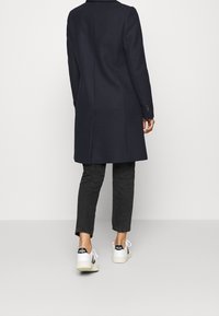 Navy wollen blendjas met een tailored fit, enkele split en knoopmanchetten, gecombineerd met zwarte denim en witte sneakers met zwarte accenten.