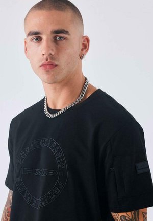 Joven con cabeza rapada, llevando un collar de cadena plateada, camiseta negra de Project X Paris y un pendiente de aro, contra un fondo claro y liso.