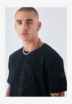 Joven con cabeza rapada, llevando un collar de cadena plateada, camiseta negra de Project X Paris y un pendiente de aro, contra un fondo claro y liso.