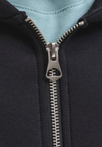 Sweat à capuche noir avec fermeture éclair argentée, doublure intérieure bleue au niveau du col, texture contrastée, et vue rapprochée de la fermeture éclair.